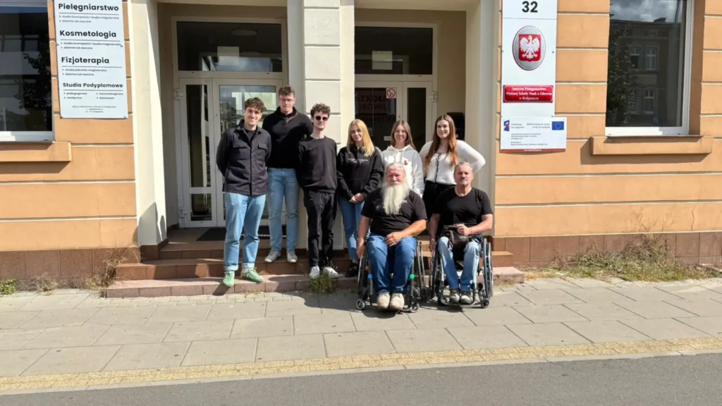 Uczestnicy szkolenia z dostępności przed budynkiem, w tym osoby z niepełnosprawnością i osoby wspierające, podczas zajęć praktycznych na zewnątrz.