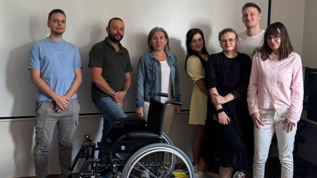 Uczestnicy szkolenia z dostępności w sali wykładowej, stojący obok wózka inwalidzkiego na tle białej tablicy.