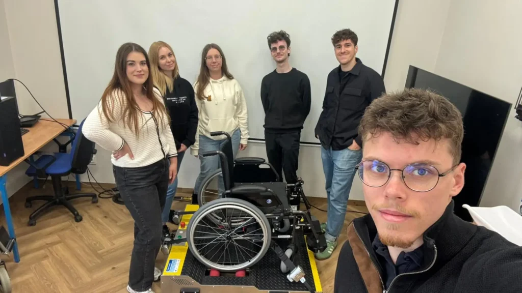 Uczestnicy szkolenia z dostępności w pracowni VR, stojący obok wózka inwalidzkiego ustawionego na specjalnym stanowisku do ćwiczeń.