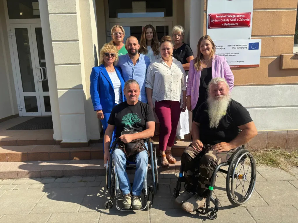 Grupa osób, w tym dziekani i prorektorzy Wyższej Szkoły Nauk o Zdrowiu oraz dwaj mężczyźni na wózkach inwalidzkich (szkoleniowcy) znajdują się przed budynkiem Instytutu Pielęgniarstwa WSNoZ.